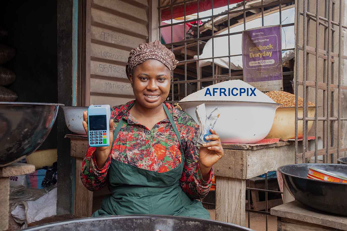 Joyful African Woman Holding Naira and POS Terminal in Front of Her Shop